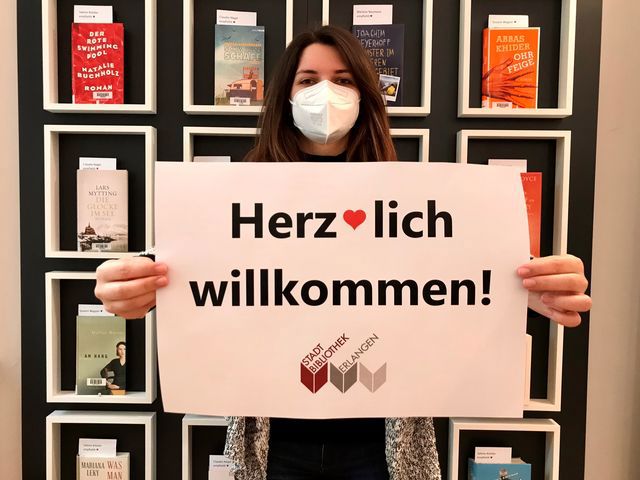 Mitarbeiterin mit Maske hält ein Schild mit Aufschrift Herzlich Willkommen